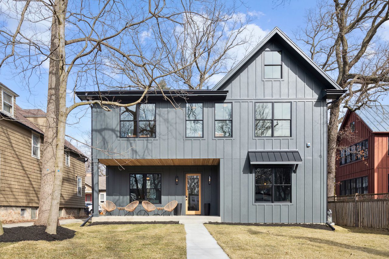 Front view of a modern two-story house with gray vertical siding, multiple windows, and a covered porch with seating. A walkway leads to the front door, and there are trees and neighboring homes in the background.